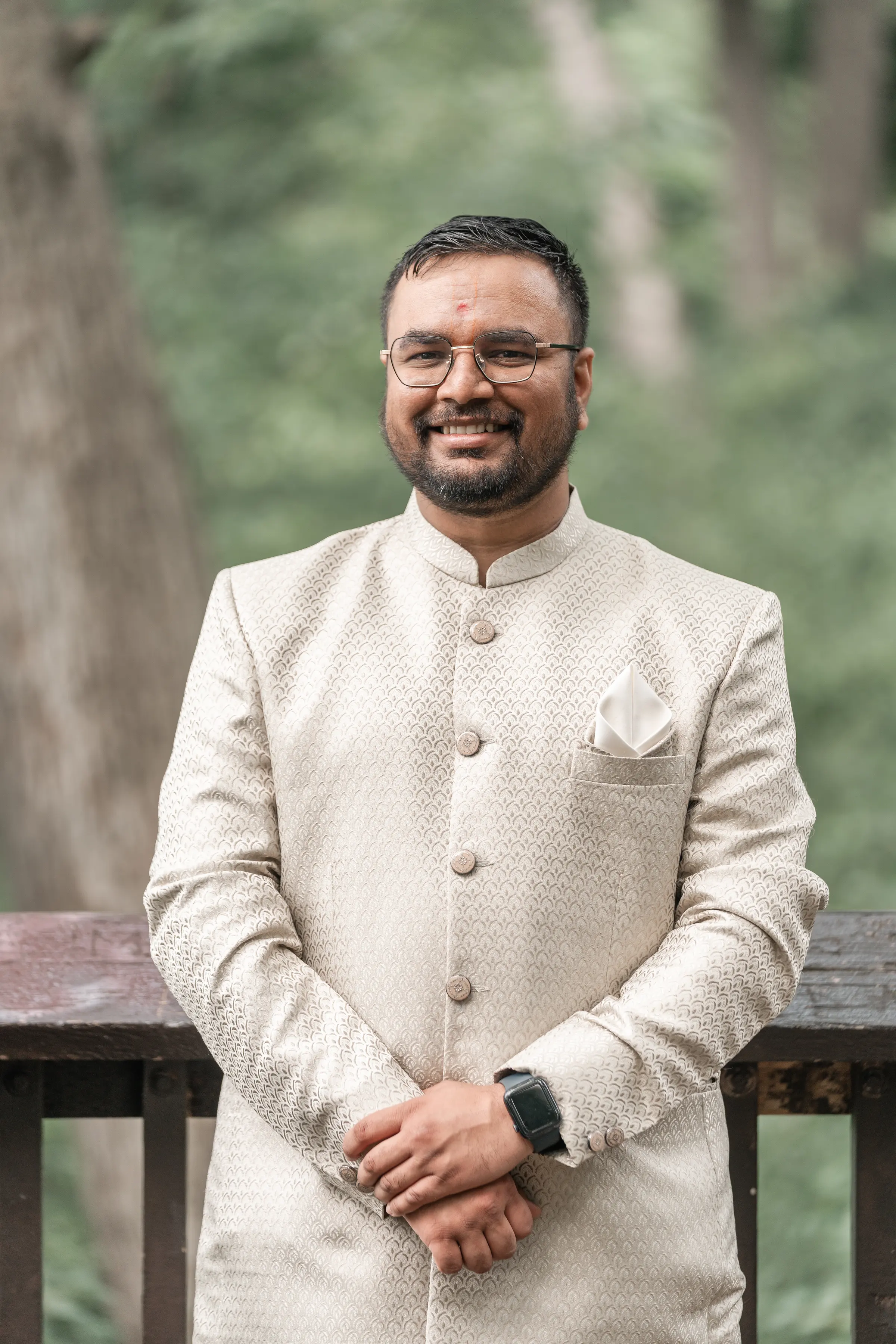 Divyesh Dobariya, in a cream sherwani, leaning against a railing with woodland behind him.