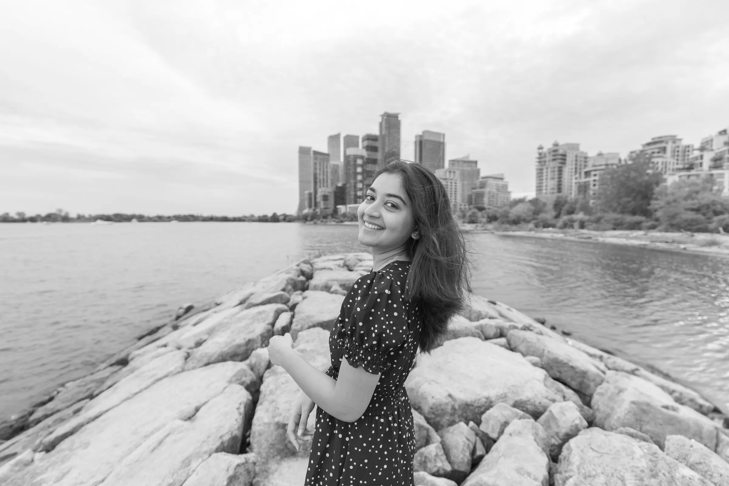 A woman on the rocks at the lakeshore, the city skyline behind her, in black and white.