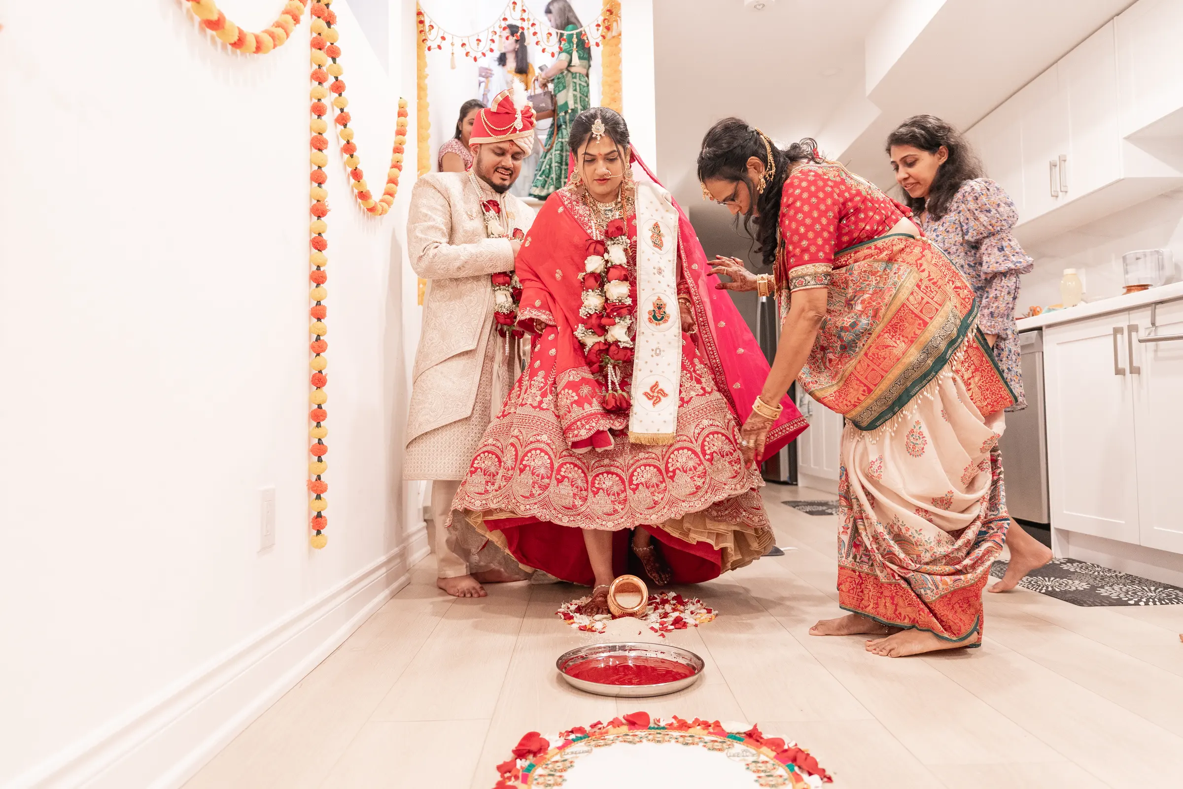 A bride is guided through the ceremony by two women, marigold garlands strung overhead.